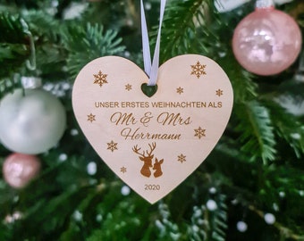 May include: A heart-shaped wooden Christmas ornament with the text "UNSER ERSTES WEIHNACHTEN ALS Mr & Mrs Herrmann 2020" and snowflake and deer designs. The ornament hangs from a white ribbon against a blurred Christmas tree background.