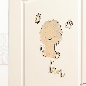 Op de afbeelding: Houten deurdecoratie met een leeuw, zon en een pootafdruk. De naam "Ian" staat onder de leeuw.