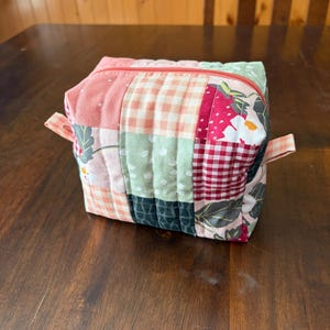 May include: A patchwork fabric zippered pouch in a cube shape. The pouch features a variety of fabrics in pink, red, green, and white, with floral and gingham patterns. The zipper is coral, and small fabric tabs are on each side.