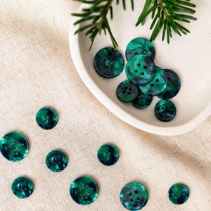 Puede incluir: Una colección de botones jaspeados verdes y negros de varios tamaños. Algunos botones están en un plato de cerámica blanca, mientras que otros están esparcidos sobre una superficie de lino beige. Una ramita de follaje verde está en el fondo.