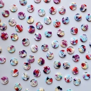 Puede incluir: Una colección de botones redondos y coloridos con un diseño veteado. Los botones presentan una mezcla de colores, incluyendo rojo, morado, azul y blanco. Cada botón tiene cuatro agujeros para coser y mide aproximadamente 2 cm de diámetro.