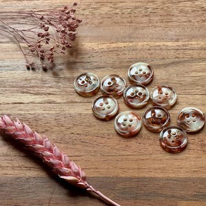 May include: A collection of ten round buttons with a marbled, tortoiseshell-like appearance. Each button has four holes and a glossy finish. The buttons are scattered on a wooden surface, with dried floral accents.