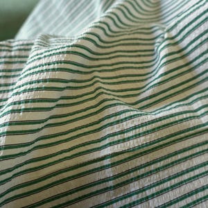 May include: Close-up of a white fabric with green and black horizontal stripes. The fabric has a textured, slightly wrinkled appearance, with the stripes running across the surface. The green stripes are evenly spaced.
