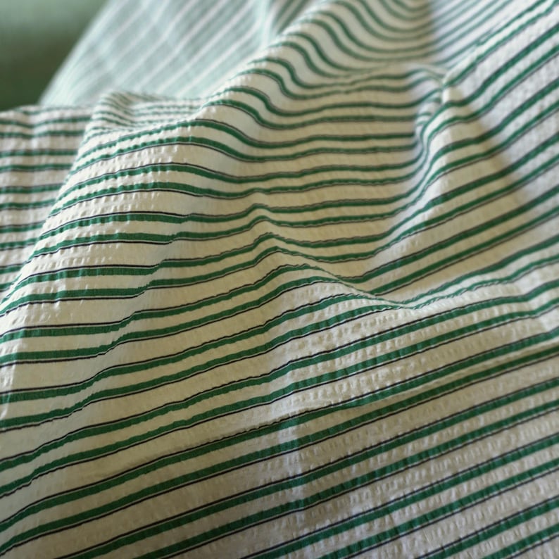 May include: Close-up of a white fabric with green and black horizontal stripes. The fabric has a textured, slightly wrinkled appearance, with the stripes running across the surface. The green stripes are evenly spaced.