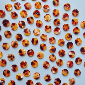 Może przedstawiać: Kolekcja okrągłych guzików z wzorem imitującym skorupę żółwia. Guziki są mieszanką kolorów bursztynowych, brązowych i kremowych, każdy z czterema otworami do szycia. Guziki są rozrzucone na jasnoniebieskiej powierzchni.