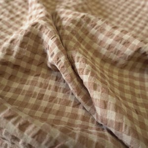 Pode incluir: Close-up de um tecido macio e enrugado com um padrão xadrez. O tecido apresenta um padrão repetitivo de pequenos quadrados castanhos claros e brancos. O material é drapeado, criando dobras e sombras que adicionam profundidade à imagem.