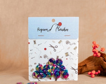 Pode incluir: Um pacote de botões coloridos e marmorizados com uma variedade de cores, incluindo azul, roxo e verde. O pacote tem um cabeçalho azul claro com o texto "Pigeon & Wishes" e um padrão de ferramentas de costura. Vários botões estão espalhados na frente do pacote.