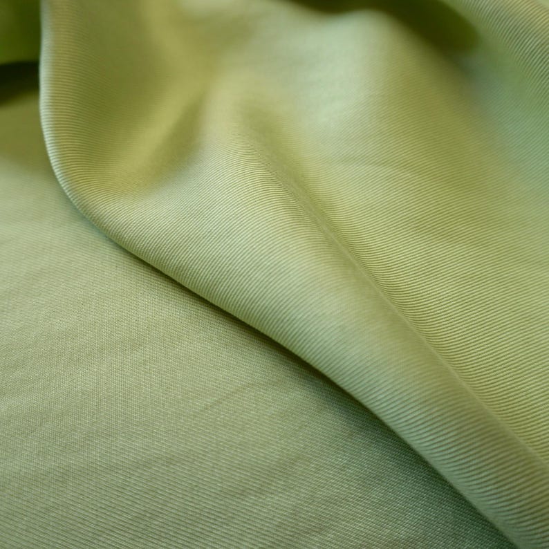 May include: Close-up of a light green fabric with a subtle ribbed texture. The fabric is draped, creating soft folds and shadows. The color is a muted, natural green, suggesting a textile suitable for clothing or home decor.