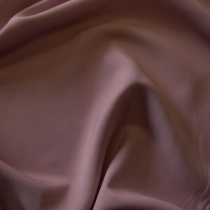 May include: Close-up of a flowing fabric in a muted mauve color. The material has a smooth, silky texture with soft folds and shadows, creating a sense of depth and movement. The fabric appears to be a luxurious textile, possibly silk or satin.