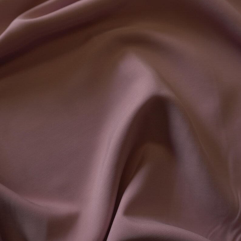May include: Close-up of a flowing fabric in a muted mauve color. The material has a smooth, silky texture with soft folds and shadows, creating a sense of depth and movement. The fabric appears to be a luxurious textile, possibly silk or satin.
