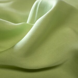 May include: Close-up of a light green fabric with a subtle ribbed texture. The fabric is draped in soft folds, creating shadows and highlights that emphasize its smooth surface. The color is a pale, muted green, reminiscent of spring foliage.