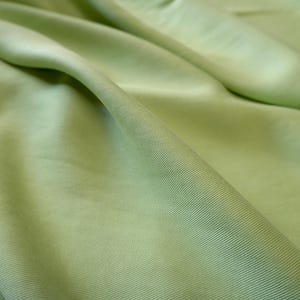 May include: Close-up of a light green fabric with subtle ribbed texture. The fabric is draped, creating soft folds and shadows. The color is a pale, muted green, reminiscent of spring foliage. The material appears smooth and possibly silky.