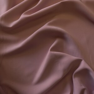 May include: Close-up of a flowing fabric in a muted mauve color. The material has a soft sheen and is draped in gentle folds, creating shadows and highlights. The fabric appears smooth and luxurious, suggesting a high-quality textile.
