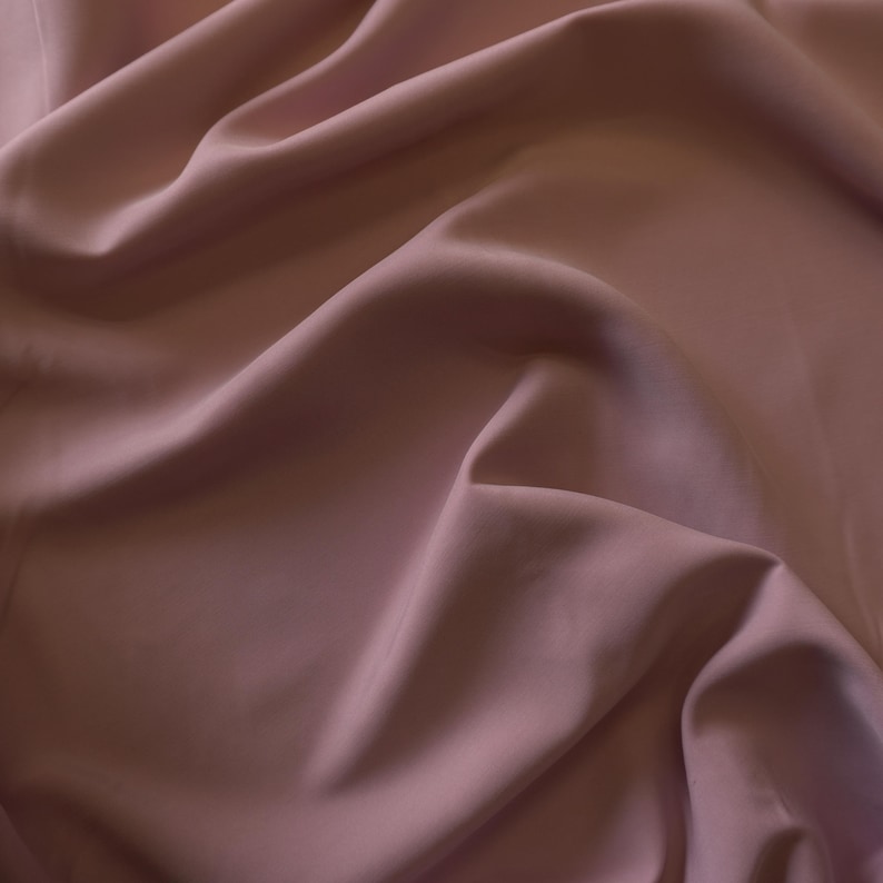 May include: Close-up of a flowing fabric in a muted mauve color. The material has a soft sheen and is draped in gentle folds, creating shadows and highlights. The fabric appears smooth and luxurious, suggesting a high-quality textile.