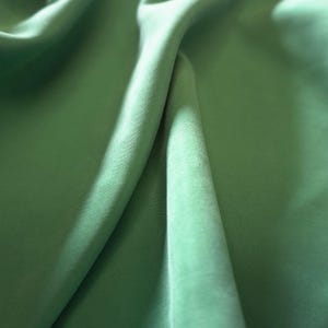 May include: Close-up of a flowing, light green fabric. The material is soft and appears to be draped, with folds and shadows creating depth. The color is a muted, natural green, suggesting a textile suitable for clothing or home decor.