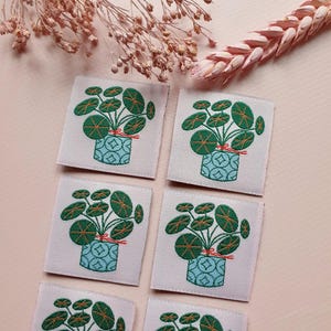 Pode incluir: Seis patches quadrados de tecido com imagens bordadas de uma planta em vaso com folhas redondas verdes. A planta est&aacute; amarrada com uma fita vermelha e o vaso &eacute; azul com um padr&atilde;o geom&eacute;trico branco. Os patches est&atilde;o sobre um fundo rosa claro.