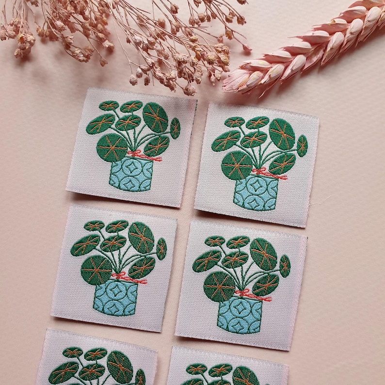 Pode incluir: Seis patches quadrados de tecido com imagens bordadas de uma planta em vaso com folhas redondas verdes. A planta est&aacute; amarrada com uma fita vermelha e o vaso &eacute; azul com um padr&atilde;o geom&eacute;trico branco. Os patches est&atilde;o sobre um fundo rosa claro.