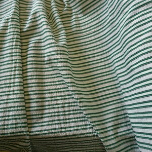 May include: A close-up shot of a fabric featuring a white base with green horizontal stripes. The fabric is folded, creating a textured effect. The stripes are evenly spaced and run across the material.