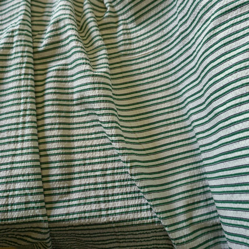 May include: A close-up shot of a fabric featuring a white base with green horizontal stripes. The fabric is folded, creating a textured effect. The stripes are evenly spaced and run across the material.
