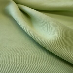 May include: Close-up of a light green fabric with a diagonal ribbed texture. The fabric is folded, creating shadows and highlighting the texture. The color is a soft, muted green, suitable for apparel or home decor.