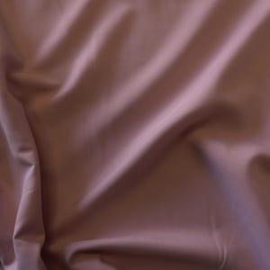 May include: A close-up shot of a fabric with a dusty rose color. The fabric is draped, creating soft folds and shadows. The material appears smooth and has a slight sheen, suggesting a luxurious texture. The overall effect is elegant and subtle.