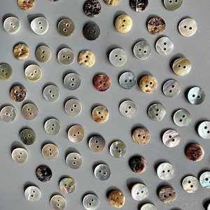 May include: A selection of round buttons in diverse colours and textures, including iridescent, brown, and grey. Each button has two or four holes for sewing. The buttons are scattered on a grey surface.