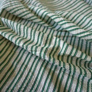 May include: A close-up shot of a white fabric featuring a green and black striped design. The material has a textured surface and a slightly creased look. The stripes are arranged vertically across the fabric, which is folded.
