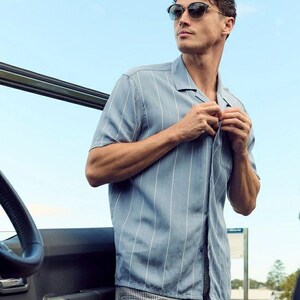 May include: A man wearing a light blue striped short-sleeved shirt with a collared neckline and a pair of gray and white checkered shorts. He is wearing sunglasses and has his hand on the button of his shirt.