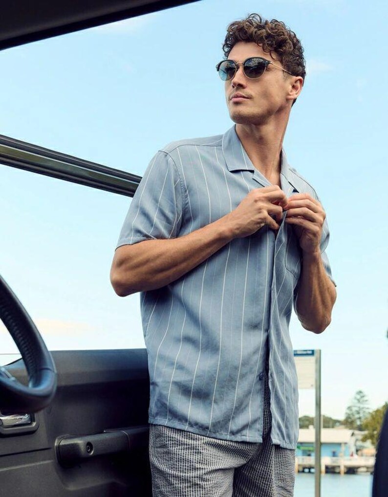 May include: A man wearing a light blue striped short-sleeved shirt with a collared neckline and a pair of gray and white checkered shorts. He is wearing sunglasses and has his hand on the button of his shirt.