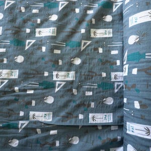 Puede incluir: Un rollo de tela con un patrón repetido de formas turquesas, blancas y marrones sobre un fondo gris. El diseño incluye palmeras, triángulos y rectángulos con el texto "NAT".