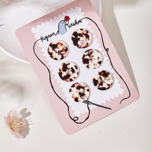 May include: A set of six round, four-hole buttons with a mottled brown and cream tortoiseshell pattern, displayed on a rectangular pink card labeled Pigeon Wishes with a graphic of a bird and a needle and thread illustration.