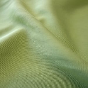 May include: Close-up of a light green fabric with a ribbed texture. The fabric is folded, creating shadows and highlights that emphasize the texture and color variations. The material has a subtle sheen.