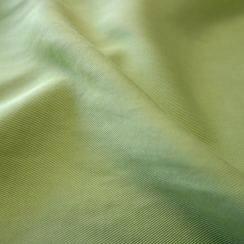 May include: Close-up of a light green fabric with a ribbed texture. The fabric is folded, creating shadows and highlights that emphasize the texture and color variations. The material has a subtle sheen.