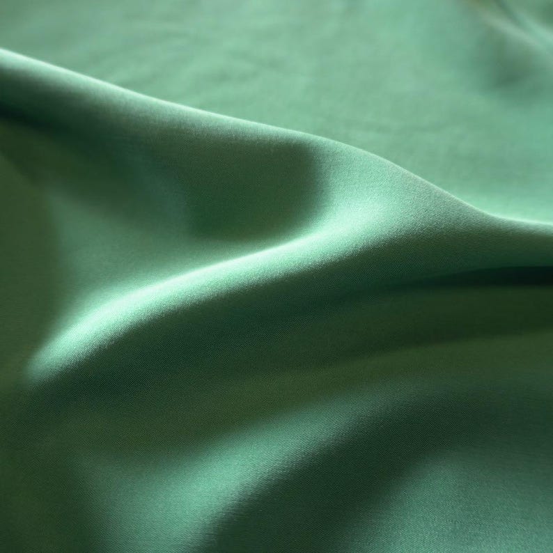 May include: Close-up of a flowing, solid green fabric. The material has a soft, matte finish with subtle folds and shadows, creating a sense of depth and texture. The color is a muted, natural green, reminiscent of foliage.