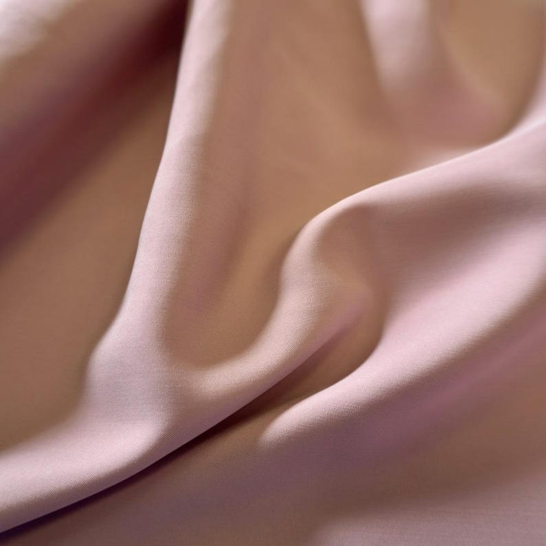 May include: Close-up of a flowing fabric in a soft, muted pink hue. The material has a smooth, silky texture with gentle folds and shadows, creating a sense of depth and movement. The overall aesthetic is elegant and delicate.