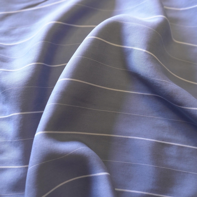 May include: A close-up of a light blue fabric with white stripes. The fabric is wrinkled and has a soft, smooth texture.