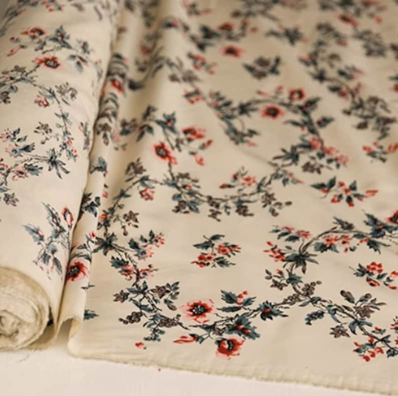 May include: Cream-colored fabric with a repeating floral pattern. The flowers are red and pink with green leaves. The fabric is rolled up and the pattern is visible on both sides.