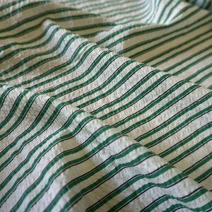May include: Close-up of a textured fabric with a repeating pattern of green and black stripes on a white background. The fabric is folded, creating shadows and depth. The stripes are evenly spaced and run diagonally across the frame.