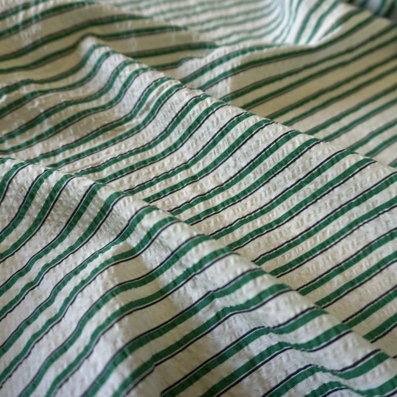 May include: Close-up of a textured fabric with a repeating pattern of green and black stripes on a white background. The fabric is folded, creating shadows and depth. The stripes are evenly spaced and run diagonally across the frame.