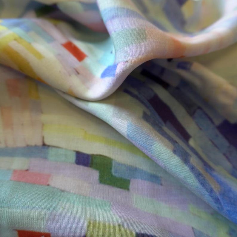 May include: Close-up of a flowing fabric with a colorful, abstract pattern. The design features rectangular shapes in shades of blue, green, yellow, pink, and purple. The fabric is soft and has a slight sheen, with visible folds and curves.