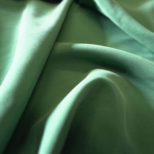 May include: Close-up of a flowing, sage green fabric. The material is smooth and appears to be a lightweight textile, with soft folds and shadows creating depth. The color is a muted, natural green.