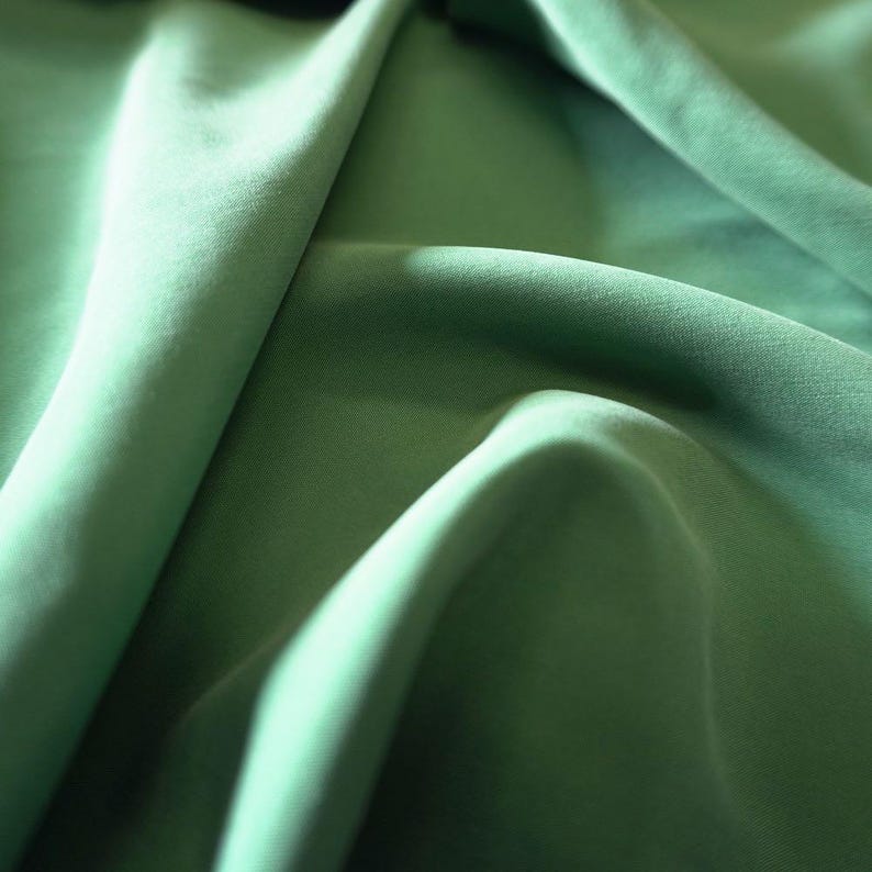 May include: Close-up of a flowing, sage green fabric. The material is smooth and appears to be a lightweight textile, with soft folds and shadows creating depth. The color is a muted, natural green.