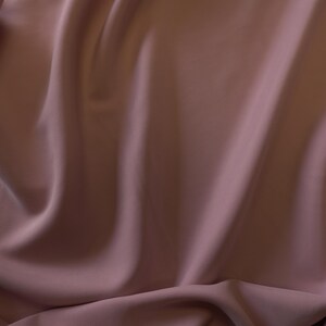May include: Close-up of a flowing fabric in a muted mauve color. The material has soft folds and a subtle sheen, creating a sense of depth and texture. The fabric appears to be smooth and lightweight, suitable for apparel or home decor.