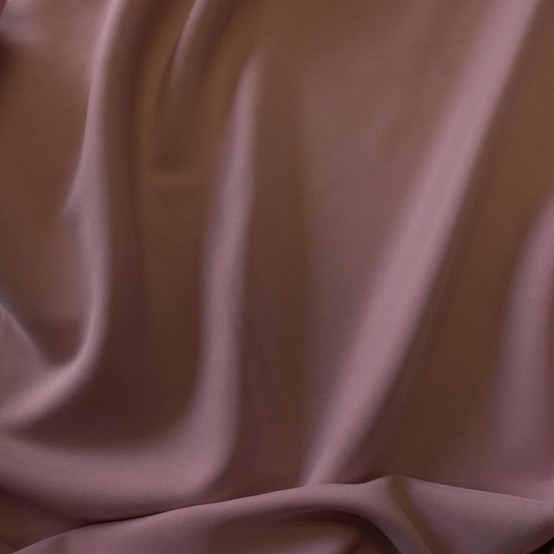May include: Close-up of a flowing fabric in a muted mauve color. The material has soft folds and a subtle sheen, creating a sense of depth and texture. The fabric appears to be smooth and lightweight, suitable for apparel or home decor.