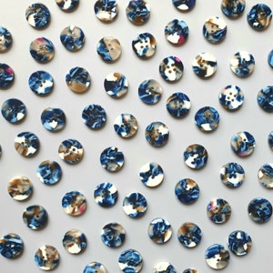Puede incluir: Colección de botones redondos con cuatro agujeros cada uno. Los botones tienen un diseño veteado en tonos azules, blancos y marrones. Los botones están dispersos sobre una superficie gris claro.
