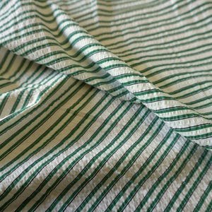 May include: Close-up of a white fabric with green and black stripes. The fabric is folded, showing the texture and pattern. The stripes are evenly spaced and run horizontally across the material, creating a classic design.