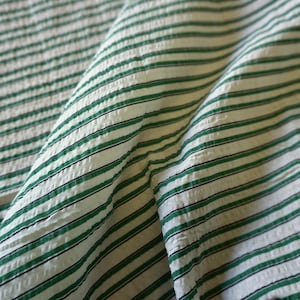 May include: Close-up of a textured fabric with a repeating pattern of green and black stripes on a white background. The fabric is folded, showing the texture and direction of the stripes. The fabric appears to be cotton or a similar material.