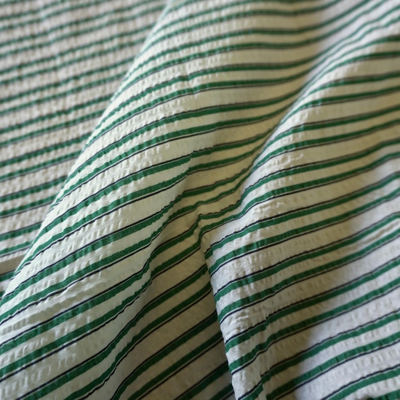 May include: Close-up of a textured fabric with a repeating pattern of green and black stripes on a white background. The fabric is folded, showing the texture and direction of the stripes. The fabric appears to be cotton or a similar material.