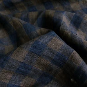 Può includere: Un tessuto intrecciato con un motivo a quadri blu scuro e marrone. Il materiale presenta una trama visibile con pieghe e increspature che creano profondità, mettendo in risalto il design a quadrati alternati sulla superficie del tessuto.