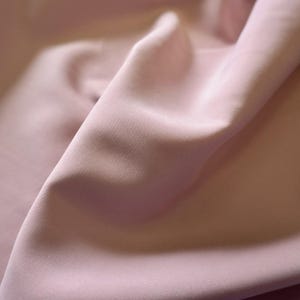May include: Close-up of a soft, flowing fabric in a muted rose color. The material is draped, creating gentle folds and shadows that highlight its texture and sheen. The fabric appears smooth and luxurious.